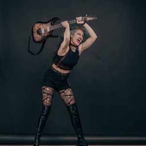 Neli Dimitrova holding Ibanez Electric Guitar, ready to smash it, with a metal outfit, harness and boots